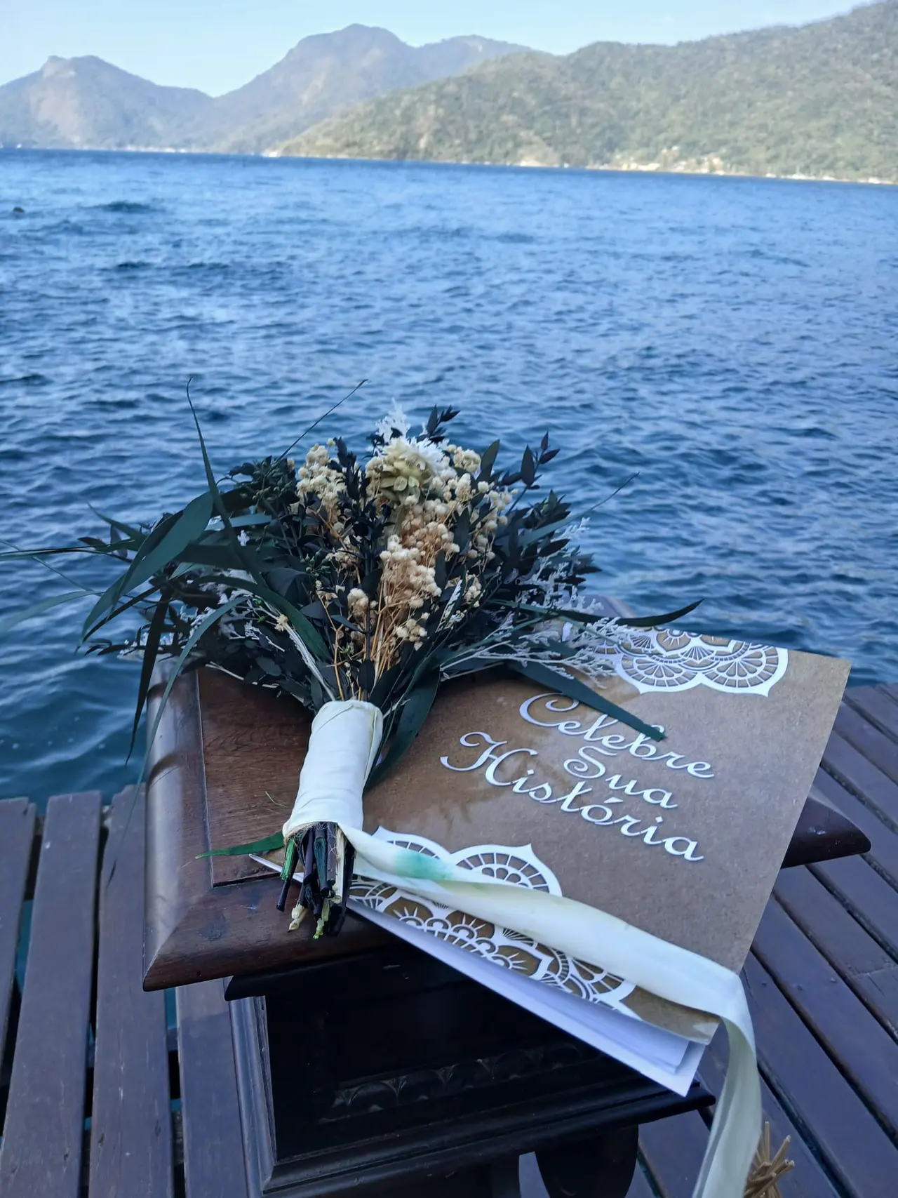 Buquê sobre o livro da cerimônia Celebre Sua História, com o mar ao fundo